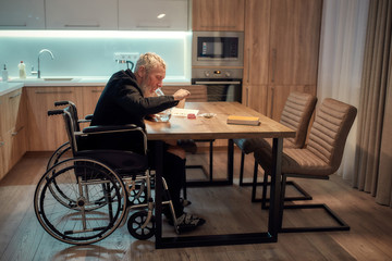 Painkiller. Senior disabled man in wheelchair lighting cannabis in the bowl of glass water pipe or bong in kitchen. Marijuana tools on the table. Legalization and dependence on light drugs concept