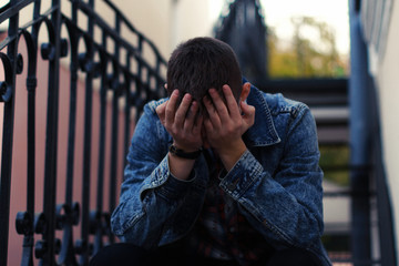 Sad young boy in jeans jacket covering face with his hands is crying and sitting on city stair.