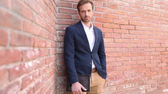 handsome elegant businessman leaning on a brick wall, holding one hand on his tablet, and the other in pocket, posing, smiling, fixing his collar