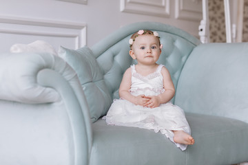 cute little girl in white dress and hoops of flowers on head is sitting on a couch in a classic style and having fun at home.