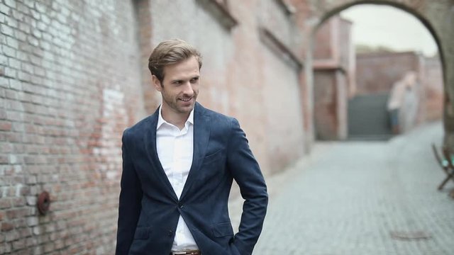 young sexy businessman walking near a brick wall, pulling out his phone from his pocket, talking on the phone and smiling