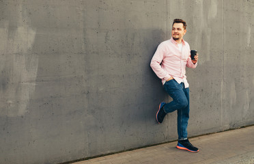 Obraz na płótnie Canvas A young handsome stylish smiling guy standing at the gray wall and drinking coffee or tea from disposable cup outdoors. City lifestyle concept