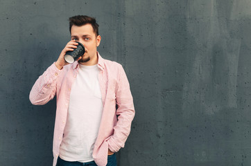 Coffee break. A young attractive charismatic man with stylish hairstyle drinking tea from disposable paper cup at the wall. Gray background with copy space