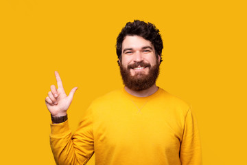 Naklejka premium Cheerful bearded man is pointing up while smiling at the camera on yellow background.