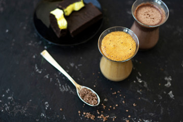 Dalgona coffee and cocoa in glass transparent glasses. Nearby lies a metal spoon with instant coffee, and a chocolate dessert on a saucer