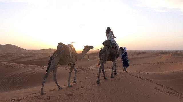 Tourist woman riding caravan of camel leading with Bedouin in blue dressing in sand dunes in Sahara desert in sunset, Morocco