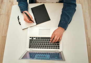 Top view of female hands working using laptop and making some notes while working remotely at home. Home office workplace. Cropped phot