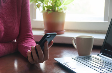 Young woman dreanking coffee while using smartphone and laptop at the kitchen table. Chatting concept. Work from home concept