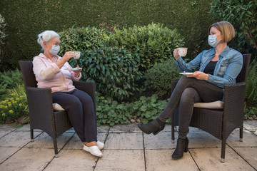 Senior woman and daughter with face masks having coffee in the garden at safeteyl distance 