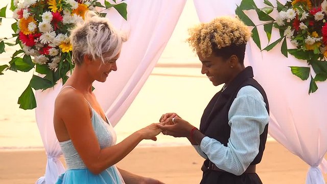 african american female groom in black suit and caucasian bride in dress in ceremony exchanging rings in hands on tropical beach under wedding flover arch