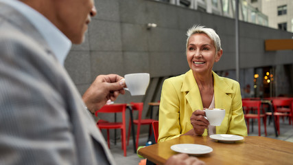 Fototapeta na wymiar Perfect morning. Beautiful and cheerful middle-aged woman in yellow suit jacket drinking fresh coffee with her friend while sitting in cafe outdoors