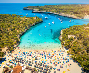 Fototapeta premium Aerial view of sandy beach with colorful umbrellas, swimming people, boats and yachts in sea bay, green trees and blue water at sunset in summer. Travel. Balearic islands, Spain. Top view. Landscape