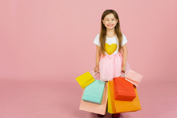 Little blonde girl enjoys her shopping on a pastel pink background with copyspace. Sale. Cute little girl with many multicolored shopping bags in studio