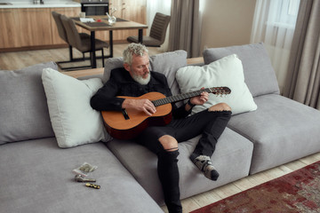 Obraz premium Let music flow. Bearded middle-aged man, artist writing song, playing guitar, sitting on the couch. Metal pipe for smoking marijuana, cannabis buds in a plastic bag and lighter lying on the couch