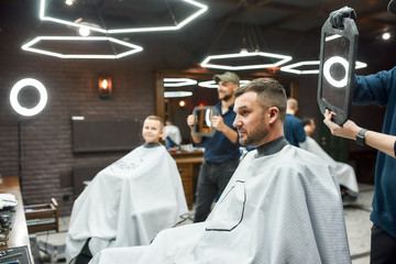 Look here. Two barbers holding mirrors and showing haircuts to father and son sitting in armchair...