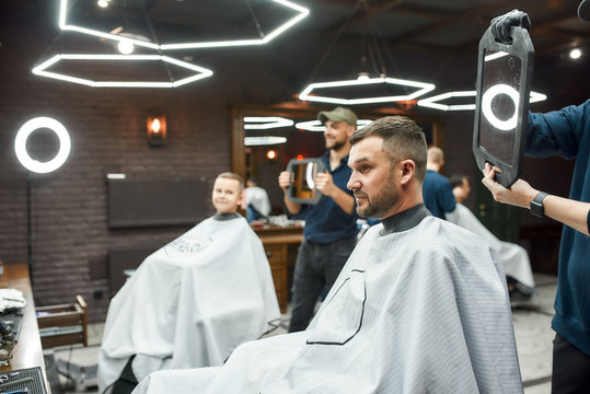 Look Here. Two Barbers Holding Mirrors And Showing Haircuts To Father And Son Sitting In Armchair At Modern Barbershop. Family Visiting Barber Shop