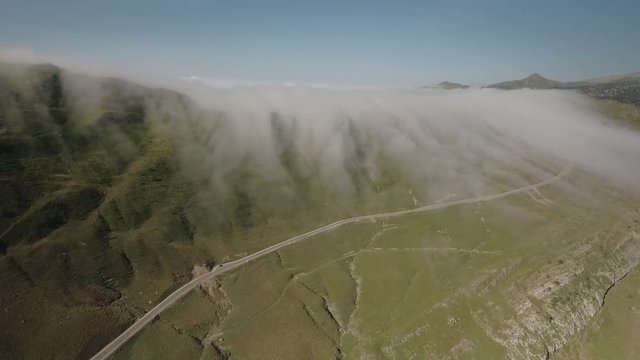AERIAL DRONE BACKWARD SMOOTH MOVEMENT OF FOG ENTERING GREEN VALLEY WITH MOUNTAIN ROAD IN THE PYRENEES, SPAIN 