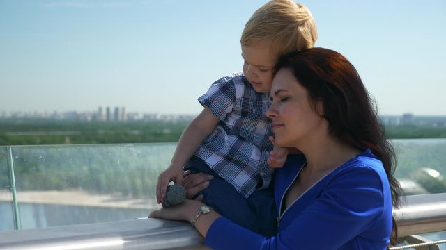 Loving Mother Hugging Caress Child While Holding on Hands on Bridge. City Background Blue Sky Bright Sunny Day. 2x Slow motion 1/2 60 FPS