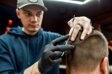 Working process. Young barber in cap with scissors in hands making haircut for his client in barber shop