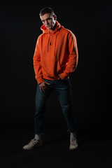 Fototapeta premium Young handsome man in jeans and orange hoodie posing while standing on a black background.