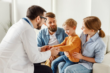 Friendly doctor is holding the hands of a child and speaking with boy and parents.