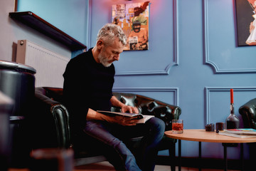 Waiting. Handsome middle-aged bearded man sitting on sofa at barbershop or salon and reading magazine