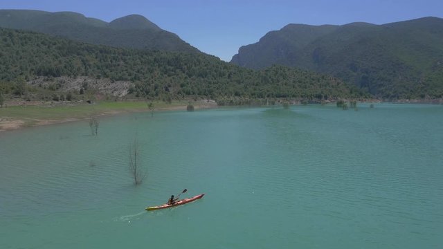 AERIAL DRONE SHOT OF ORANGE CANOE AND MAN ALONE IN A TURQUOISE RESERVOIR AND STUNNING MOUNTAIN VALLEY