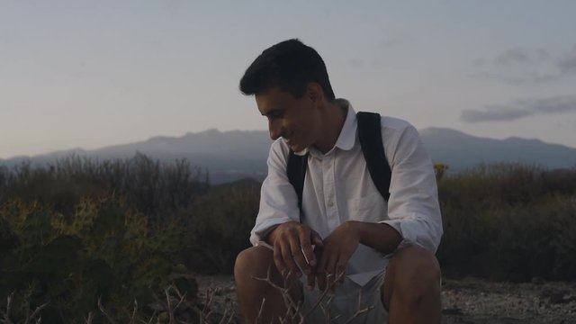 A traveler with a backpack in the mountains on an island in white sits in the mountains near a cactus and touches it.