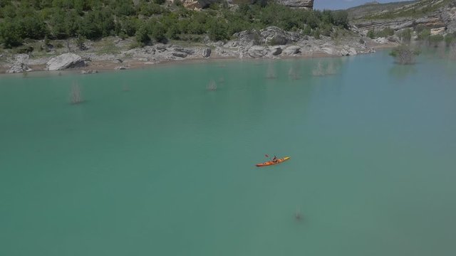 AERIAL DRONE SHOT OF ORANGE CANOE AND MAN ALONE IN A TURQUOISE RESERVOIR, TILT UP DISCOVERING VALLEY. 