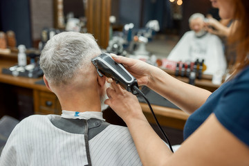Perfect work. Barber girl using hair clipper. Making new haircut for elegant mature man sitting in armchair in the front of mirror