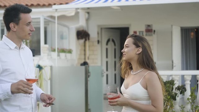 A couple in white against the background of the villa drinks rose wine . The guy gives the girl a cigar, the girl coughs and refuses. Cigars, the harm of smoking. Girl coughs from cigars