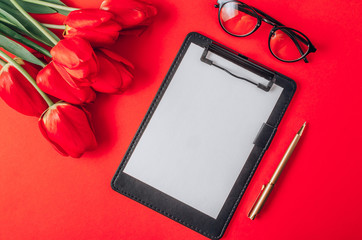 Home office workplace: clipboard, eyeglasses, keyboard, tulip flowers on red background. Top view, copy space