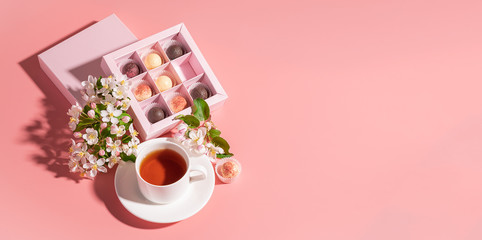 Chocolates of various colors in a festive elegant box packing a cup of tea on pastel pink background decoration flowers.