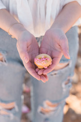 pink mini donut in the hands of a young woman in white blouse and ripped jeans
