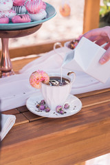 Hand pouring milk in a cup and saucer of  coffee on a feminine outdoor wood table with a side donut and edible dried rose buds