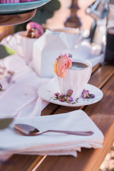 Romantic cup of coffee with pink donut on an outdoor wood  table with feminine dried rosebuds on saucer 