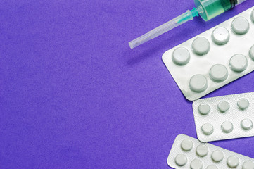 Three shiny packets of white pills and a medical syringe with a closed needle on a blue background. Diseases, pharmaceuticals.