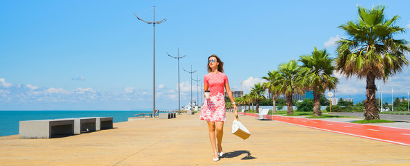 Young beautiful woman in red dress walking on the beach boulevard with coffee cup and bag. Banner...