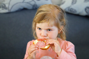 Adorable little toddler girl eating fresh prepared salami sandwith in kitchen. Happy child eats healthy food for lunch or dinner. Baby learning. Home, nursery, playschool or daycare