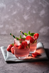 Fresh strawberry lemonade with ice, mint and paper straw in sparkling glasses on gray table background, copy space. Cold summer drink. Berry cocktail