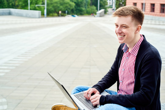 Happy smiling student sitting in university campus and working with laptop outdoors, copy space. Preparing for exam, typing, surfing internet. Technology, education concept