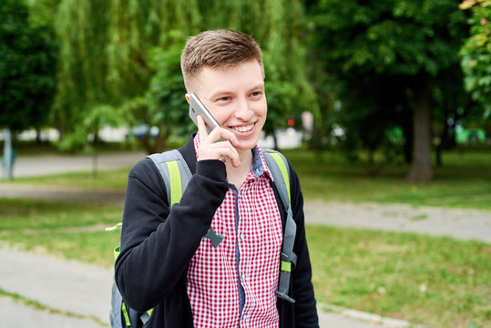 Portrait of happy smiling young student with backpack walking in park and tolking on mobile cellphone outdoors, copy space