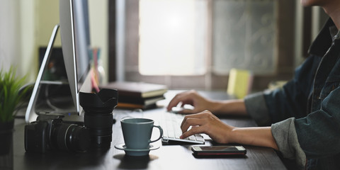 Cropped image of photographer hands typing on wireless keyboard and using mouse while sitting in front computer monitor that putting on working desk.