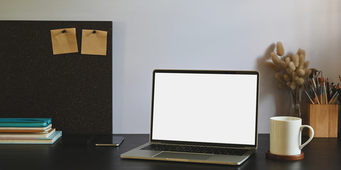 Photo of white blank screen computer laptop putting on black working desk and surrounded by stack of notebook, smartphone, pencil holder, wild grass in vase and coffee cup.