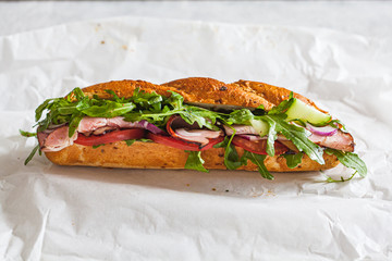 Baguette sandwich with meat, tomato, cucumber and arugula on white paper. Take away food concept.