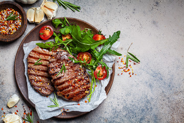Grilled beef steaks with arugula and tomato salad on wooden plate, dark background, copy space, top view.