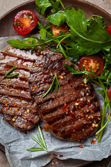 Grilled beef steaks with arugula and tomato salad on wooden plate, dark background, top view.