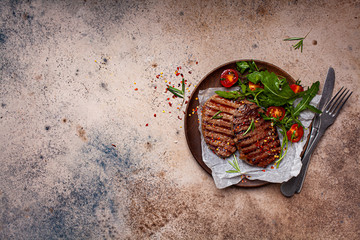 Grilled beef steaks with arugula and tomato salad on wooden plate, dark background, copy space, top view.