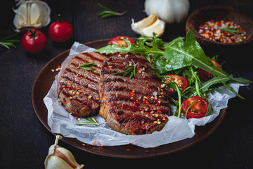 Grilled beef steaks with arugula and tomato salad on wooden plate, dark background.
