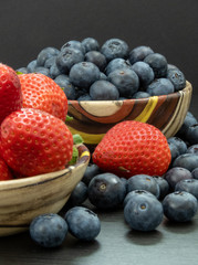 Blueberries and Strawberries in ceramic bowls and on a black wooden table. Healthy red berry snack. Fresh vegan breakfast. Close up. Bunch of vitamins.